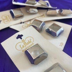 May include: Silver-toned, geometric buttons from La Mode Buttons are displayed on white cards against a purple background. The buttons have a shiny, metallic finish and a faceted design. Each button measures 1.25 inches.