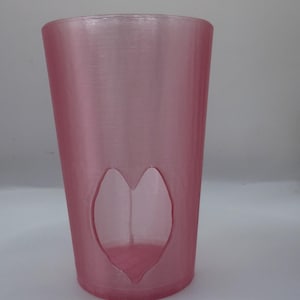 May include: A pink, cylindrical glass with a heart-shaped cutout. The glass has a smooth, translucent appearance and is likely made of plastic or glass. The heart cutout adds a decorative element to the glass.