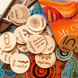 May include: Wooden discs with etched designs and words like "Sandhill Cloud," "Kangaroo," and "People Sitting" are displayed. They are arranged on a vibrant, patterned surface beside a book titled "My Co." The image has a warm, earthy colour palette.
