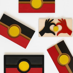 May include: Wooden wall art pieces in the Aboriginal flag design. The art features rectangular and angled shapes with black, red, yellow, and brown colours. One piece depicts hands forming a heart around a yellow circle.