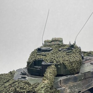 Puede incluir: Maqueta detallada de un tanque militar verde oliva, con red de camuflaje. El tanque tiene un cañón largo, múltiples ruedas y dos antenas delgadas. La red de camuflaje es una mezcla de verde y marrón, cubriendo partes del tanque.