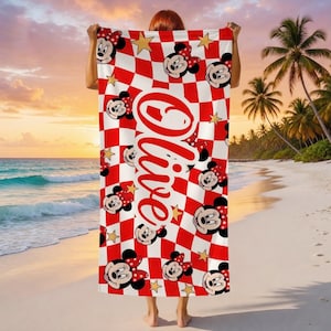 Peut inclure: Une serviette de plage &agrave; carreaux rouges et blancs avec des visages de Minnie Mouse et des &eacute;toiles dor&eacute;es. La serviette porte le nom "Olive" en &eacute;criture rouge. La serviette est tenue sur un fond de plage.