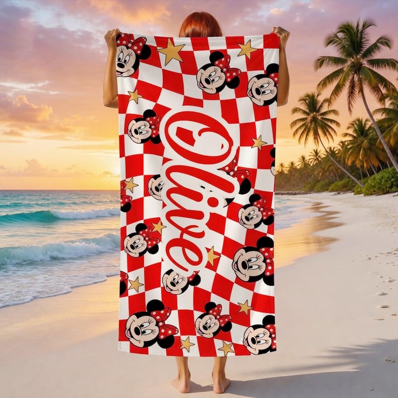 Peut inclure: Une serviette de plage &agrave; carreaux rouges et blancs avec des visages de Minnie Mouse et des &eacute;toiles dor&eacute;es. La serviette porte le nom "Olive" en &eacute;criture rouge. La serviette est tenue sur un fond de plage.