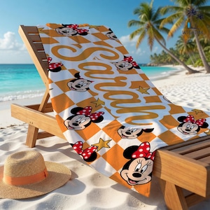 Peut inclure: Serviette de plage &agrave; carreaux orange et blanc avec des visages de Minnie Mouse et le nom "Sarah". Le tissu est pos&eacute; sur une chaise longue en bois sur une plage de sable.