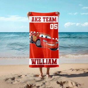 May include: Red beach towel with a cartoon race car design. The towel has the text "AKZ TEAM 05" at the top and "WILLIAM" at the bottom. The background shows a beach and the sea.
