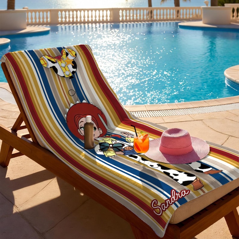 Puede incluir: Una toalla de playa a rayas de colores con un dise&ntilde;o de personaje de dibujos animados est&aacute; colocada en una tumbona de madera. Un sombrero para el sol rosa, gafas de sol y una bebida est&aacute;n sobre la toalla. El nombre "Sandra" est&aacute; escrito en la toalla.