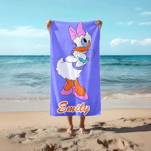 K&ouml;nnte beinhalten: Ein lavendelfarbenes Strandtuch mit der Zeichentrickfigur Daisy Duck. Das Handtuch zeigt ein gro&szlig;es Bild von Daisy Duck mit einer rosa Schleife, Schuhen und einem blauen Armband. Der Name "Emily" steht unten in orangefarbener Schrift.