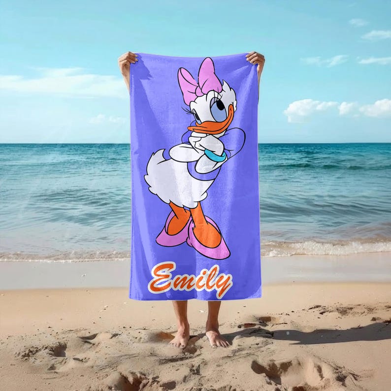 K&ouml;nnte beinhalten: Ein lavendelfarbenes Strandtuch mit der Zeichentrickfigur Daisy Duck. Das Handtuch zeigt ein gro&szlig;es Bild von Daisy Duck mit einer rosa Schleife, Schuhen und einem blauen Armband. Der Name "Emily" steht unten in orangefarbener Schrift.