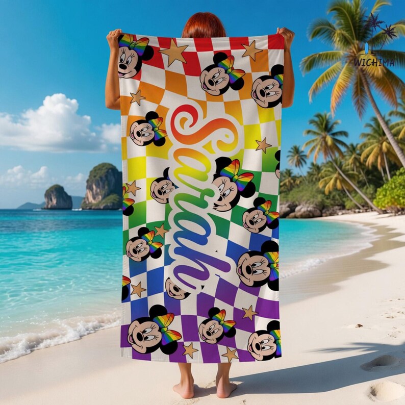 Peut inclure: Une serviette de plage avec un motif &agrave; carreaux aux couleurs de l'arc-en-ciel, des visages de Minnie Mouse et des &eacute;toiles dor&eacute;es. Le nom "Sarah" est &eacute;crit verticalement en d&eacute;grad&eacute; arc-en-ciel. La serviette est tenue sur une plage.