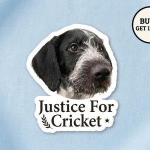 May include: A sticker featuring a dog's head with black and white fur, and the text "Justice For Cricket". A circular graphic in the upper right corner reads "BUY 5 GET 1 FREE". The sticker is on a light blue background.