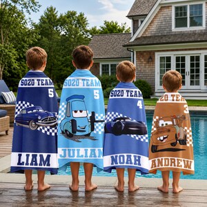 May include: Four children stand near a swimming pool, each wearing a personalised towel. The towels are blue and brown, featuring cartoon car designs, names, and the text "SOZO TEAM" and numbers. The towels have a checkered flag pattern.