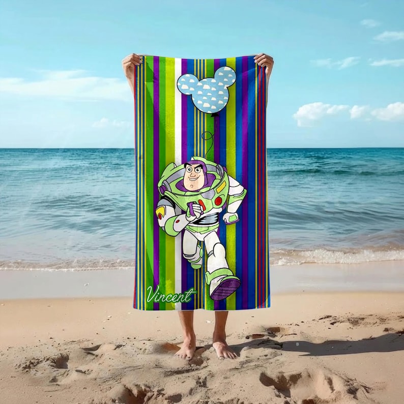 Puede incluir: Toalla de playa colorida con una imagen de dibujos animados de Buzz Lightyear corriendo, con un dise&ntilde;o de cabeza de Mickey Mouse. La toalla tiene rayas verticales en verde, morado y azul. El nombre "Vincent" est&aacute; impreso.