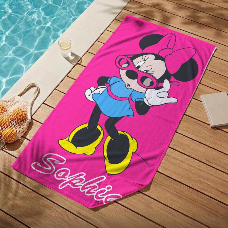 K&ouml;nnte beinhalten: Ein leuchtend pinkfarbenes Strandtuch mit einem Cartoon-Design von Minnie Mouse, die eine Sonnenbrille und ein blaues Kleid tr&auml;gt. Der Name "Sophie" ist in wei&szlig;er Schrift auf dem Handtuch zu sehen. Es liegt auf einem Holzdeck in der N&auml;he eines Pools.