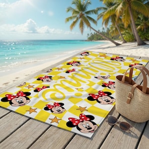 Peut inclure: Serviette de plage &agrave; carreaux jaunes et blancs avec des visages de Minnie Mouse et des &eacute;toiles. La serviette porte le nom "Minnie" en &eacute;criture cursive. La serviette est sur une terrasse en bois pr&egrave;s de la plage.
