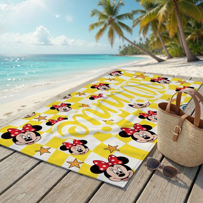 Peut inclure: Serviette de plage &agrave; carreaux jaunes et blancs avec des visages de Minnie Mouse et des &eacute;toiles. La serviette porte le nom "Minnie" en &eacute;criture cursive. La serviette est sur une terrasse en bois pr&egrave;s de la plage.