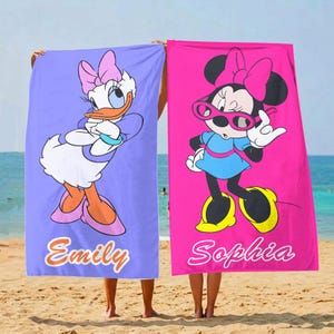 K&ouml;nnte beinhalten: Zwei Strandt&uuml;cher mit Disney-Figuren. Eines ist lavendelfarben mit Daisy Duck, das andere pink mit Minnie Mouse. Auf jedem Handtuch ist ein Name aufgedruckt: "Emily" und "Sophia". Die Handt&uuml;cher werden an einem Sandstrand hochgehalten.