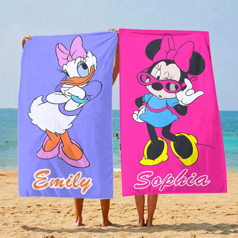 K&ouml;nnte beinhalten: Zwei Strandt&uuml;cher mit Disney-Figuren. Eines ist lavendelfarben mit Daisy Duck, das andere pink mit Minnie Mouse. Auf jedem Handtuch ist ein Name aufgedruckt: "Emily" und "Sophia". Die Handt&uuml;cher werden an einem Sandstrand hochgehalten.