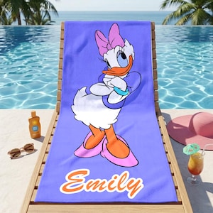 K&ouml;nnte beinhalten: Ein lila Strandtuch mit der Cartoonfigur Daisy Duck. Das Handtuch hat den Namen "Emily" in orangefarbener Schrift. Das Handtuch liegt auf einer Holzliege in der N&auml;he eines Pools, mit Sonnenbrille, Sonnencreme und einem Cocktail.