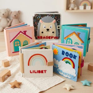 May include: A collection of handmade felt quiet books featuring various designs including a hedgehog, houses, a rainbow with the name LILIBET, and a blue book cover with clouds, a sun, a rainbow, and the text BOOK ME NAYOTDB.