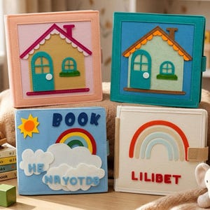 May include: Four square felt books with colorful appliqués. Two books feature house designs with doors and windows. One book displays a sun, rainbow, clouds, and the text BOOK HE NAYOTDB. One book shows a rainbow and the name LILIBET in red letters.