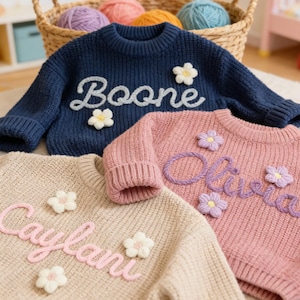 May include: Three knitted sweaters in navy, pink, and beige, each with a name embroidered in a contrasting colour. The navy sweater says "Boone", the pink sweater says "Olivia", and the beige sweater says "Caylani". Each sweater has flower embellishments.
