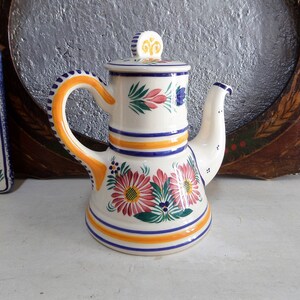 May include: A white ceramic teapot with a blue and yellow floral design. The teapot has a round body with a flared spout and a handle. The lid has a knob in the shape of a flower.