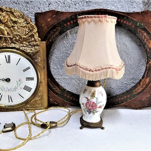 Puede incluir: Una lámpara de porcelana vintage con un diseño floral y una pantalla blanca con ribete rosa. La lámpara está sentada en una mesa frente a un marco de madera con un diseño pintado en oro.