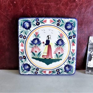Può includere: Una piastrella quadrata in ceramica con un designo floreale blu e bianco. Il centro della piastrella presenta una donna con un vestito giallo e rosso che tiene un fiore. La piastrella è circondata da un bordo di fiori blu e bianchi.
