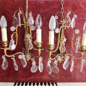 Könnte beinhalten: Eine goldfarbene Wandleuchte mit fünf Kerzenhaltern und Kristalltropfen. Die Leuchte hat ein florales Design und ist mit kleinen, dekorativen Blumen verziert.