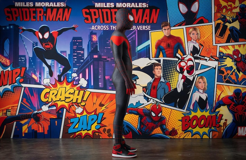 May include: A person in a black and red Spider-Man costume stands in front of a comic book-style backdrop. The backdrop features the words "Miles Morales: Spider-Man Across the Spider-Verse" and comic book sound effects like "Crash!" and "Zap!".