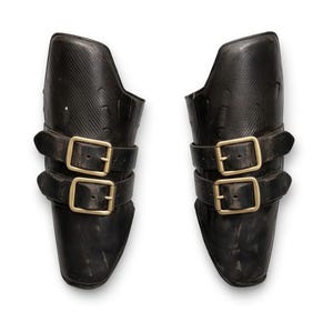 May include: A pair of black leather arm guards, each featuring two gold-coloured buckle straps. The guards are designed to fit the forearm, possibly for protection or costume use. The background is plain white.