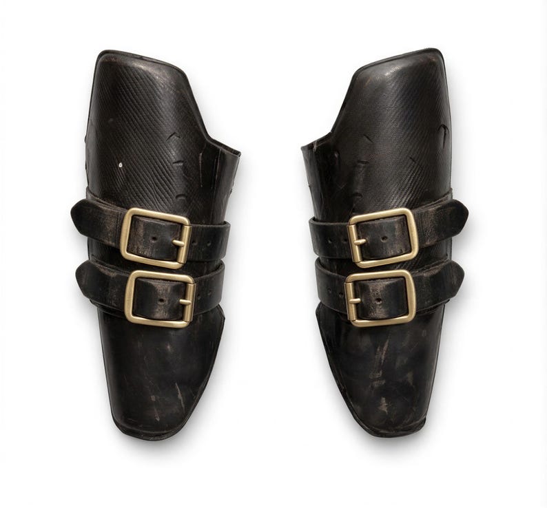 May include: A pair of black leather arm guards, each featuring two gold-coloured buckle straps. The guards are designed to fit the forearm, possibly for protection or costume use. The background is plain white.