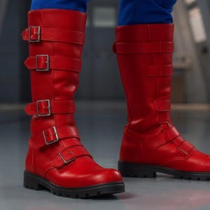 K&ouml;nnte beinhalten: Knallrote kniehohe Stiefel mit silbernen Schnallen und schwarzen Sohlen. Die Stiefel sind aus glattem Material gefertigt. Die Stiefel werden mit einer blauen Hose getragen. Die Stiefel sind ca. 40 cm hoch.