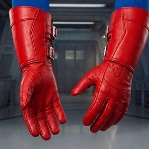 K&ouml;nnte beinhalten: Ein Paar leuchtend rote Lederhandschuhe mit langen Manschetten und silbernen Schnallen. Die Handschuhe weisen Gebrauchsspuren auf, mit sichtbaren Knitterfalten. Sie scheinen Teil eines Kost&uuml;ms oder einer Uniform zu sein. Der Hintergrund ist ein verschwommener Innenraum mit industriellen Elementen.