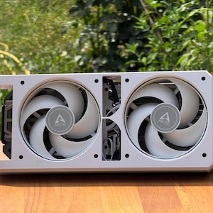 May include: A white graphics card featuring two large cooling fans with the Arctic logo centred on each. The rectangular housing has a clean, matte white finish with visible black internal heat sink fins and a metallic connector bracket on the side.