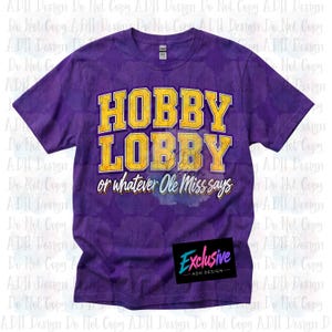 May include: A purple short-sleeve t-shirt featuring the yellow text HOBBY LOBBY in a bold collegiate font, followed by the white cursive text or whatever Ole Miss says. The shirt has a crew neckline and a standard fit with a tag at the collar.