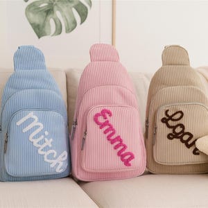 Op de afbeelding: Drie corduroy slingrugzakken in lichtblauw, roze en beige, elk met een naam geborduurd op de voorzak. Een stervormig kussen met een lachend gezicht staat naast de beige rugzak. Een olifantenbeeldje staat links.