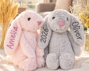 May include: Two plush bunny rabbits, one pink and one gray, are displayed. The pink bunny has the name "Amelia" embroidered on its ear. The gray bunny has the name "Oliver" and the number "2026" embroidered on its ears. Both bunnies have pink noses.