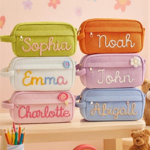 May include: A selection of corduroy pencil cases in various colours, each embroidered with a name: Sophia, Noah, Emma, John, Charlotte, and Abigail. The cases are green, orange, white, pink, and blue, with decorative embellishments.