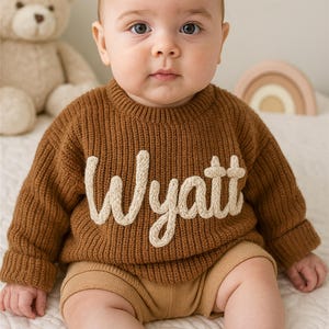 May include: A baby in a brown knitted sweater with the name "Wyatt" in cream-coloured script. The baby wears brown shorts and sits on a white surface. A teddy bear and a wooden rainbow are in the background.