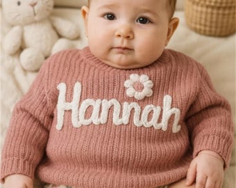 May include: A baby wearing a dusty rose-colored knit sweater with the name "Hannah" in white script. A white flower applique is on the sweater. The baby is also wearing floral print diaper covers. A stuffed bunny is in the background.