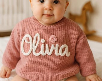 May include: A baby wearing a dusty rose-colored sweater with the name "Olivia" in white script. A small flower is next to the name. The baby is also wearing a floral diaper.