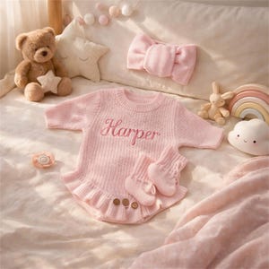 Könnte beinhalten: Ein rosa Baby-Outfit-Set mit dem Namen "Harper" auf der Vorderseite gestickt. Das Set enthält einen Strampler, Socken und ein passendes Stirnband. Ein Teddybär, ein Sternkissen und anderes Babyzubehör sind ebenfalls im Bild.
