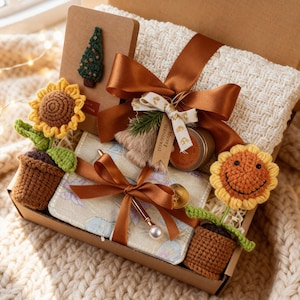 May include: A gift box containing a cream blanket, a notebook with a green tree, a small jar, a pen, and a floral wallet. The box is decorated with brown ribbons and crochet sunflowers.