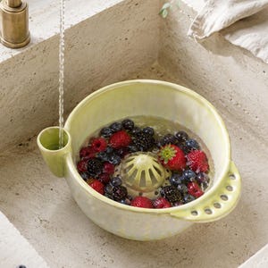 Puede incluir: Un colador de bayas de cerámica de color amarillo pálido con una cúpula de drenaje central, un pico lateral para la entrada de agua y un asa con cinco perforaciones circulares. El cuenco contiene agua, fresas rojas, arándanos y moras.