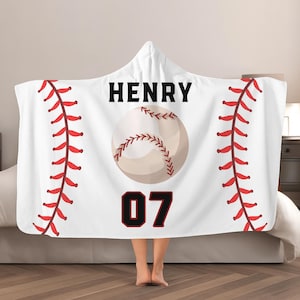 May include: White hooded blanket with a baseball design. The blanket features a baseball graphic, the name "HENRY" in black, and the number "07" in black with a red outline. Red stitching lines the sides.