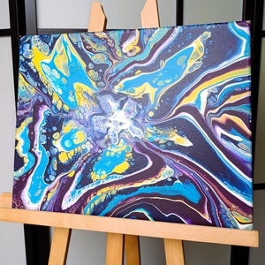 K&ouml;nnte beinhalten: Abstrakte Acrylgussmalerei auf Leinwand, ausgestellt auf einer h&ouml;lzernen Staffelei. Das Kunstwerk zeigt eine lebendige Mischung aus Farben, darunter Blau, Gelb und Lila, die ein dynamisches, flie&szlig;endes Design erzeugt. Die Leinwand ist ca. 30 cm x 30 cm gro&szlig;.