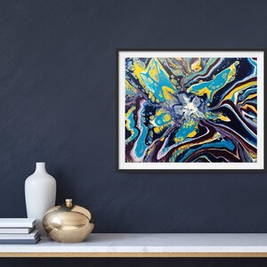 K&ouml;nnte beinhalten: Abstrakter Kunstdruck in einem schwarzen Rahmen. Das Kunstwerk zeigt ein lebendiges, farbenfrohes Design mit Blau-, Gelb- und Lilat&ouml;nen. Das Gem&auml;lde wird auf einem wei&szlig;en Tisch mit goldenem Sockel zusammen mit dekorativen Gegenst&auml;nden ausgestellt.