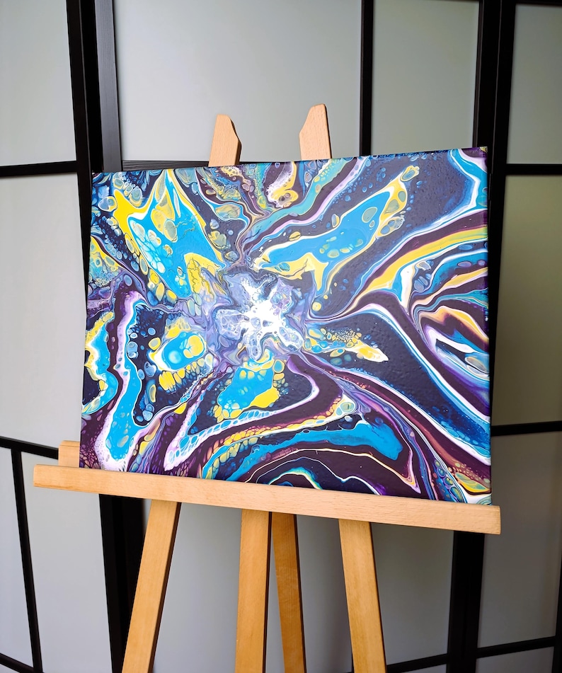 K&ouml;nnte beinhalten: Abstrakte Acrylgussmalerei auf Leinwand, ausgestellt auf einer h&ouml;lzernen Staffelei. Das Kunstwerk zeigt eine lebendige Mischung aus Farben, darunter Blau, Gelb und Lila, die ein dynamisches, flie&szlig;endes Design erzeugt. Die Leinwand ist ca. 30 cm x 30 cm gro&szlig;.