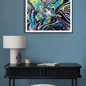 K&ouml;nnte beinhalten: Abstrakter Kunstdruck in einem schwarzen Rahmen. Das Kunstwerk zeigt ein lebendiges, flie&szlig;endes Design mit Wirbeln aus Gelb, Blau, Wei&szlig; und Lila. Der Druck h&auml;ngt &uuml;ber einem schwarzen Schreibtisch mit Lampe, Statue und Buch.